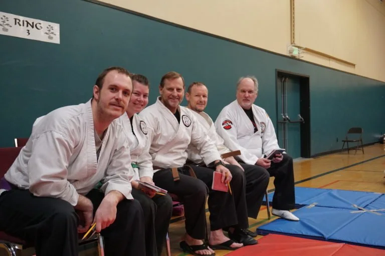 Five men in martial arts uniforms sitting on chairs in a gymnasium, holding notebooks and pens.