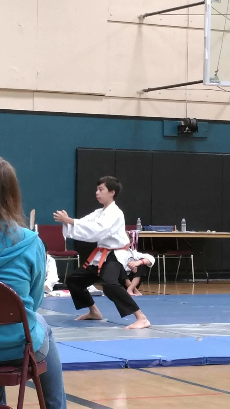 A young boy in a white martial arts uniform practicing martial arts in a gym, with an orange belt, on a blue mat during a competition or demonstration.