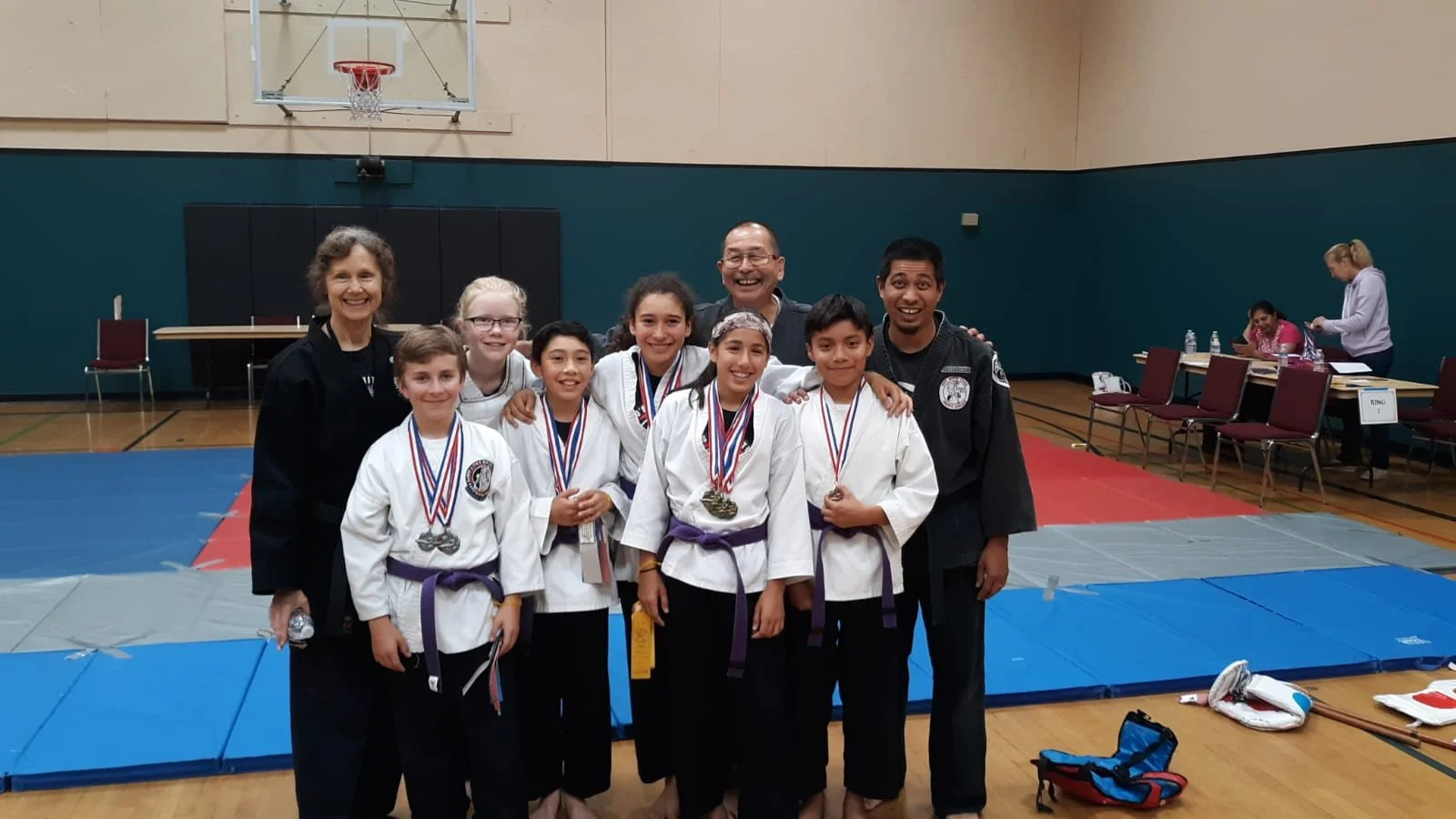 Group of young martial artists with medals, posing with their coaches or instructors in a gymnasium after a competition.