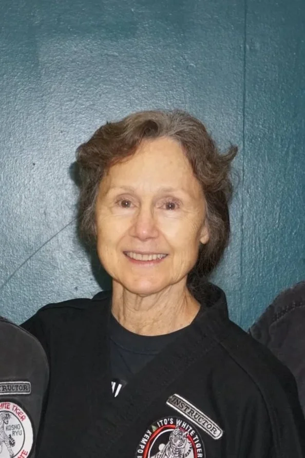 Smiling elderly woman with short gray hair wearing a black martial arts uniform with patches, standing against a blue wall.
