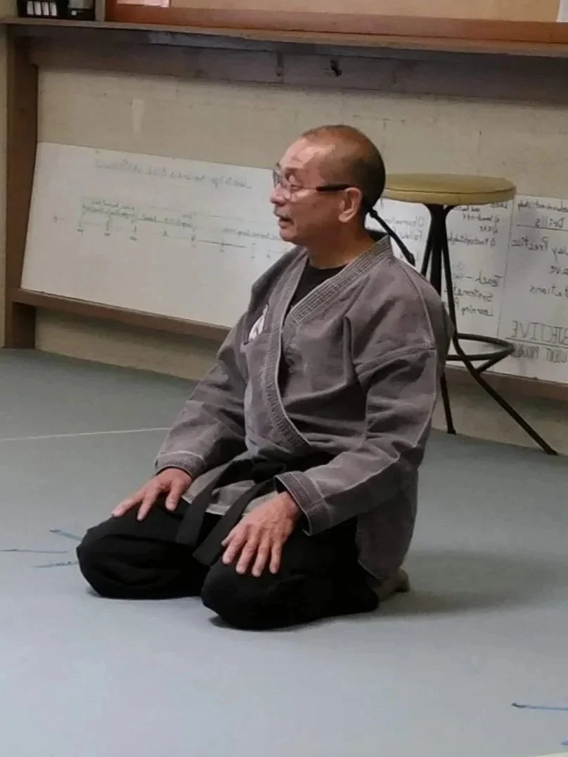 A man in a martial arts uniform kneeling on a mat, possibly in a dojo or training room, with handwritten notes on the whiteboard behind him.