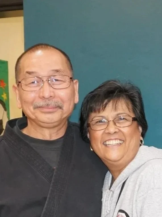 A smiling elderly man with glasses and a woman with glasses, smiling, standing close together indoors against a blue wall.
