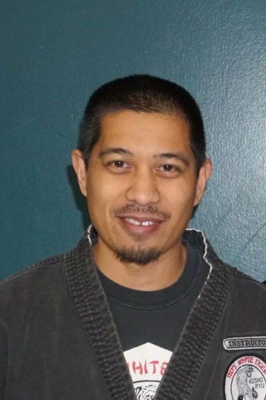 A man with short dark hair and a goatee smiling, wearing a black martial arts gi with a patch, against a blue wall.