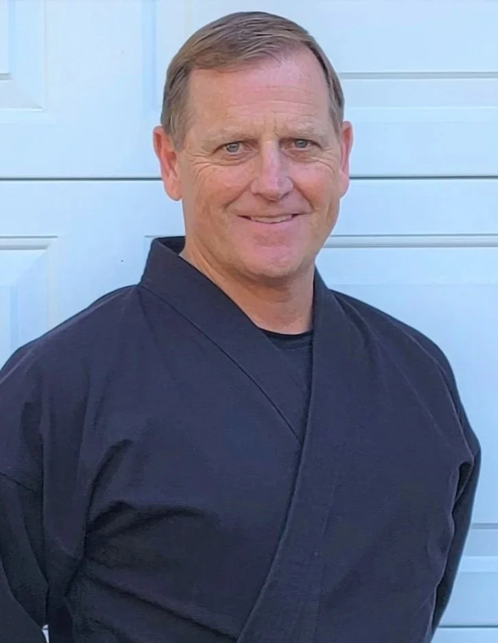 A man with short, light brown hair and blue eyes smiling, wearing a dark martial arts uniform, standing in front of a white garage door.