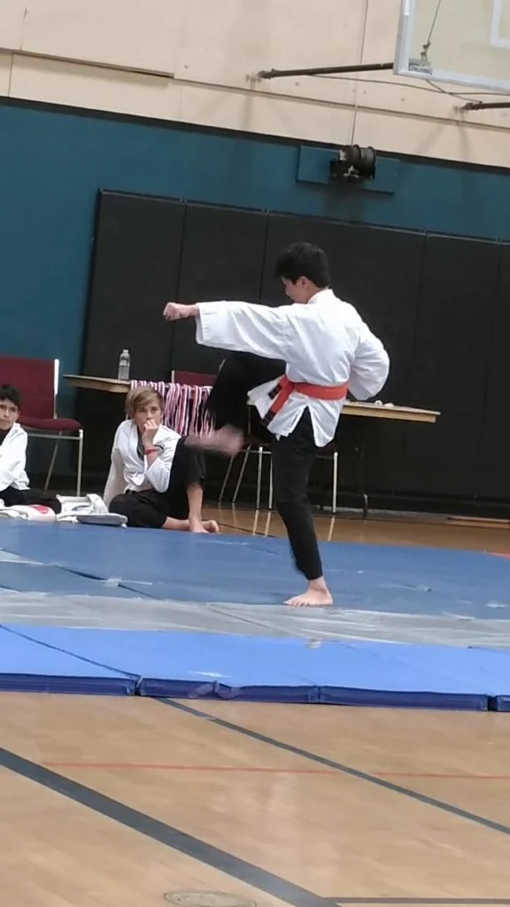 A martial arts demonstration in a gym, with a young man in a white karate gi and orange belt executing a kick on a blue mat, while two children sit nearby watching.