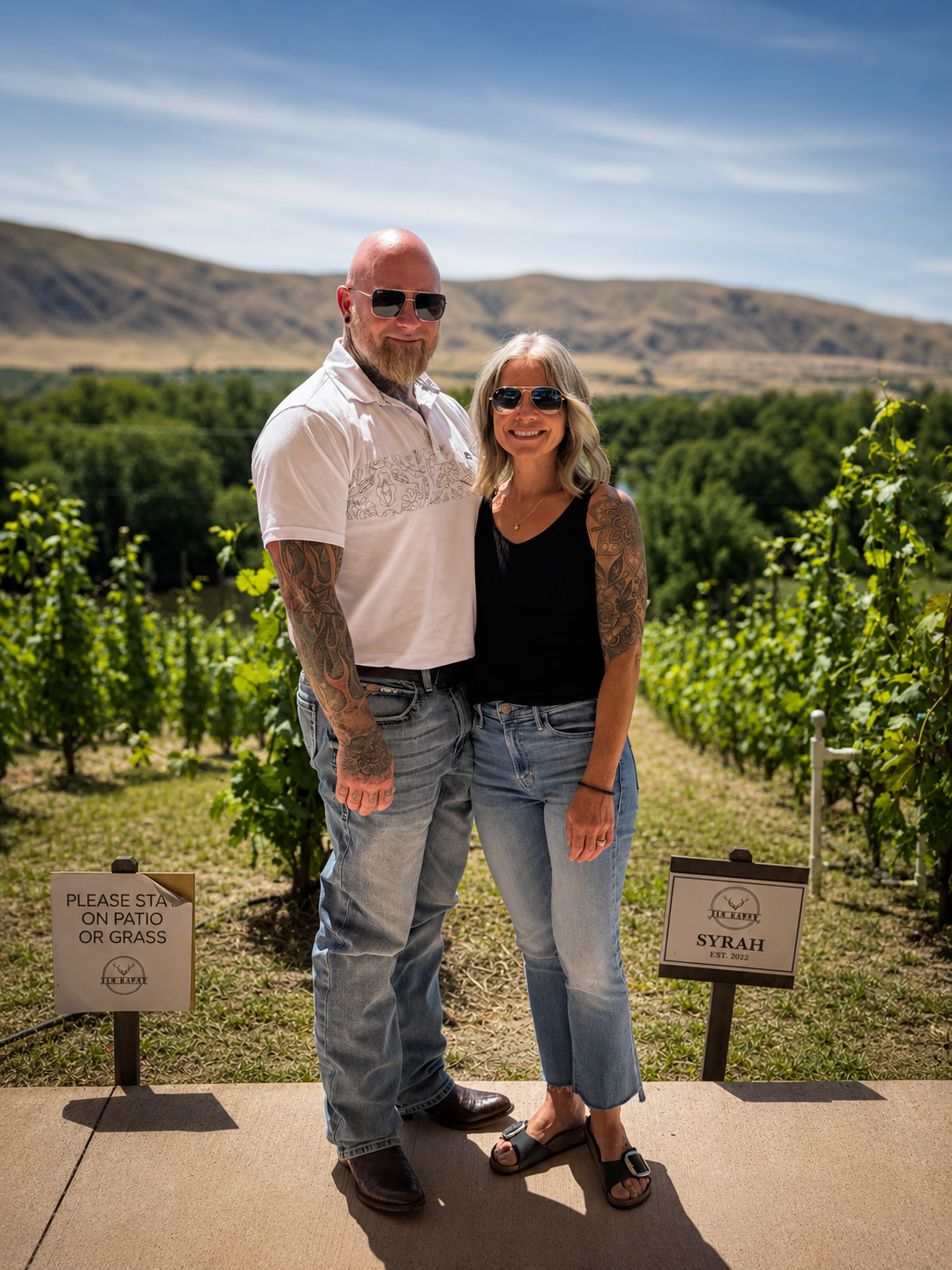 A couple standing together outdoors in a vineyard, smiling and wearing sunglasses, with mountains in the background. The man has a bald head, beard, tattoos on arms, and is wearing a white polo shirt and jeans. The woman has blonde hair, tattoos on arms, and is wearing a black top and jeans.