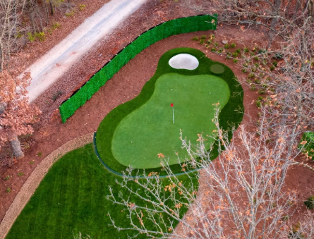 Aerial view of a golf course green with a sand trap, surrounded by trees and a dirt path.