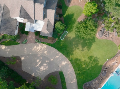 Aerial view of a backyard with a house, a curved concrete pathway, a grassy area, trees, and a small pool.