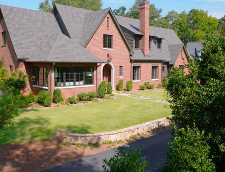 Large brick house with a steep roof and chimney, surrounded by a well-manicured lawn and bushes, with a driveway in the foreground and trees in the background.