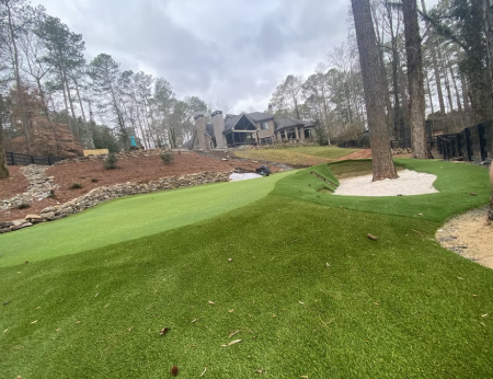View of a golf course with a putting green, sand bunker on the right, and a wooded area in the background under cloudy sky.