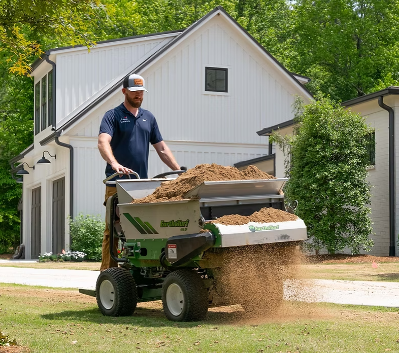 A man using a soil screener machine to separate soil and debris in a residential yard with a large white house and green trees in the background.