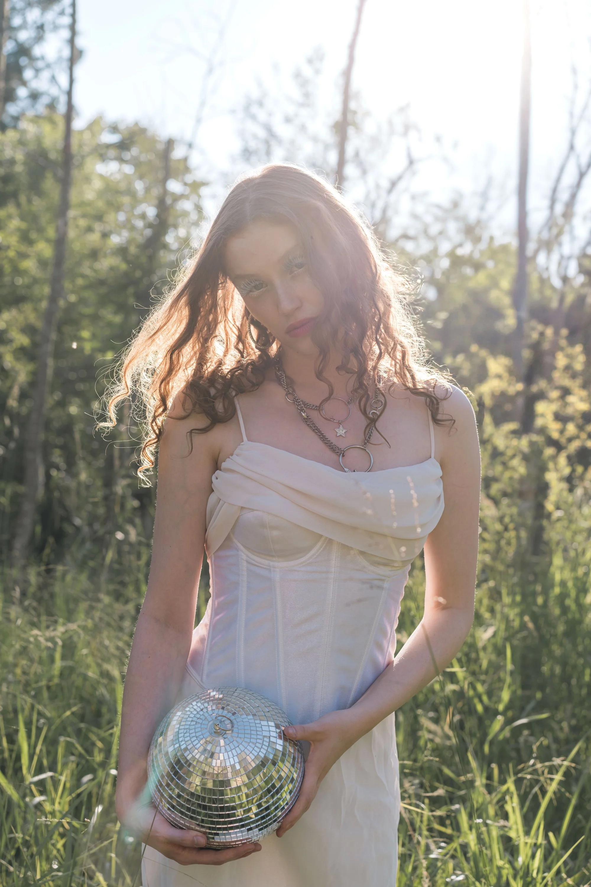 Woman with red curly hair holding a disco ball in a sunlit forest with light reflections across her face, ethereal fine art portrait photography by Missoula Montana photographer