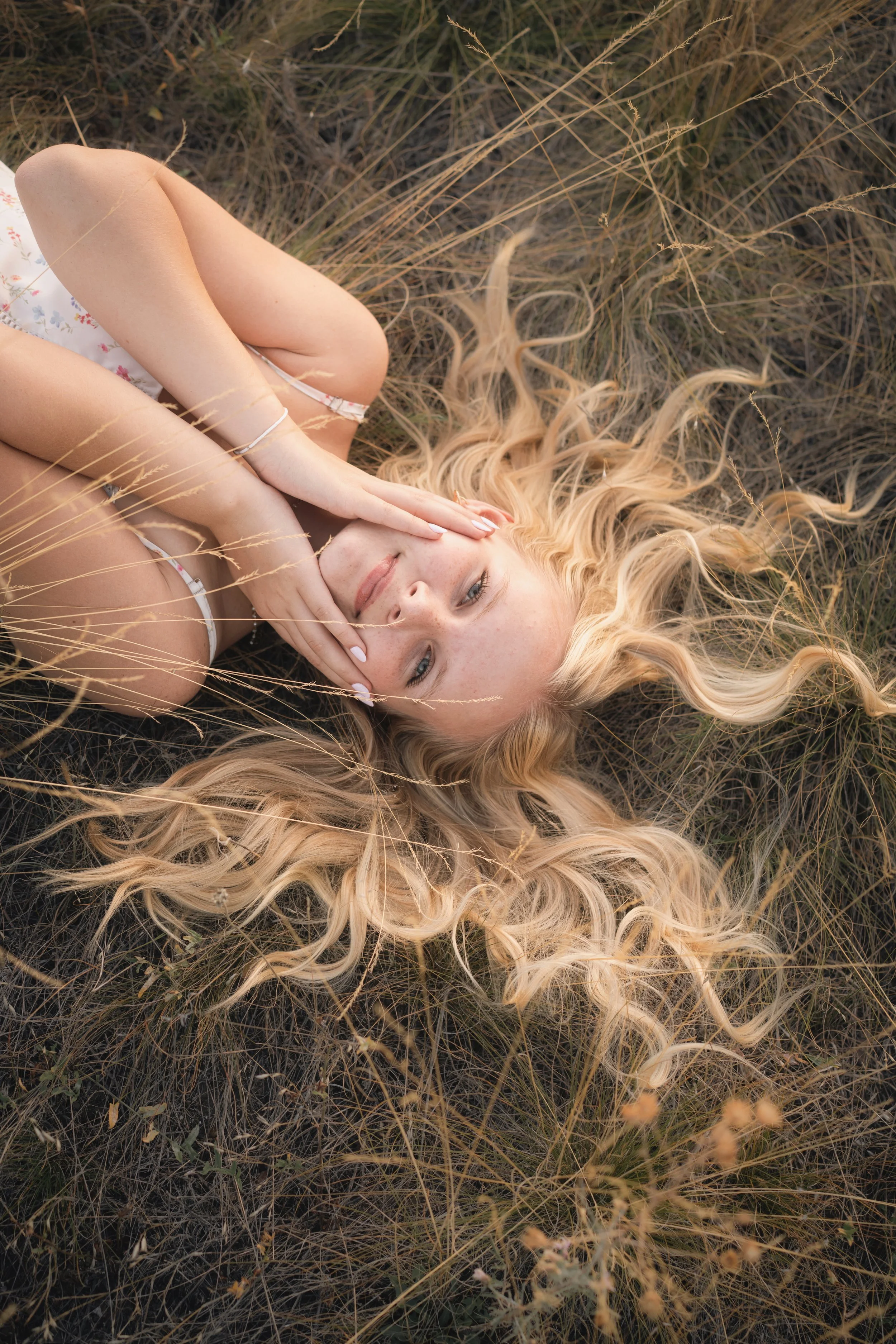 Woman lying in dry grass with long blonde curly hair spread around her and hands cradling her face, intimate fine art portrait photography by Missoula Montana photographer.