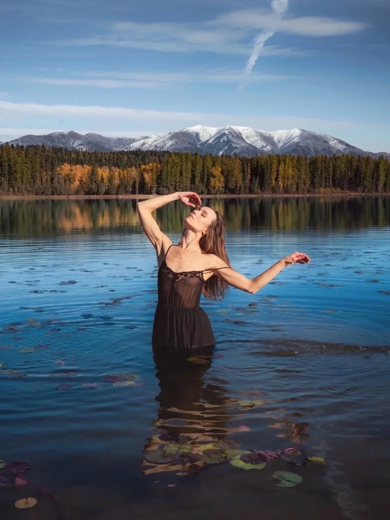 Self portrait of woman in black dress standing in Seeley Lake Montana with snow capped mountains and autumn forest reflected behind her, wild nature photography by Missoula Montana photographer.