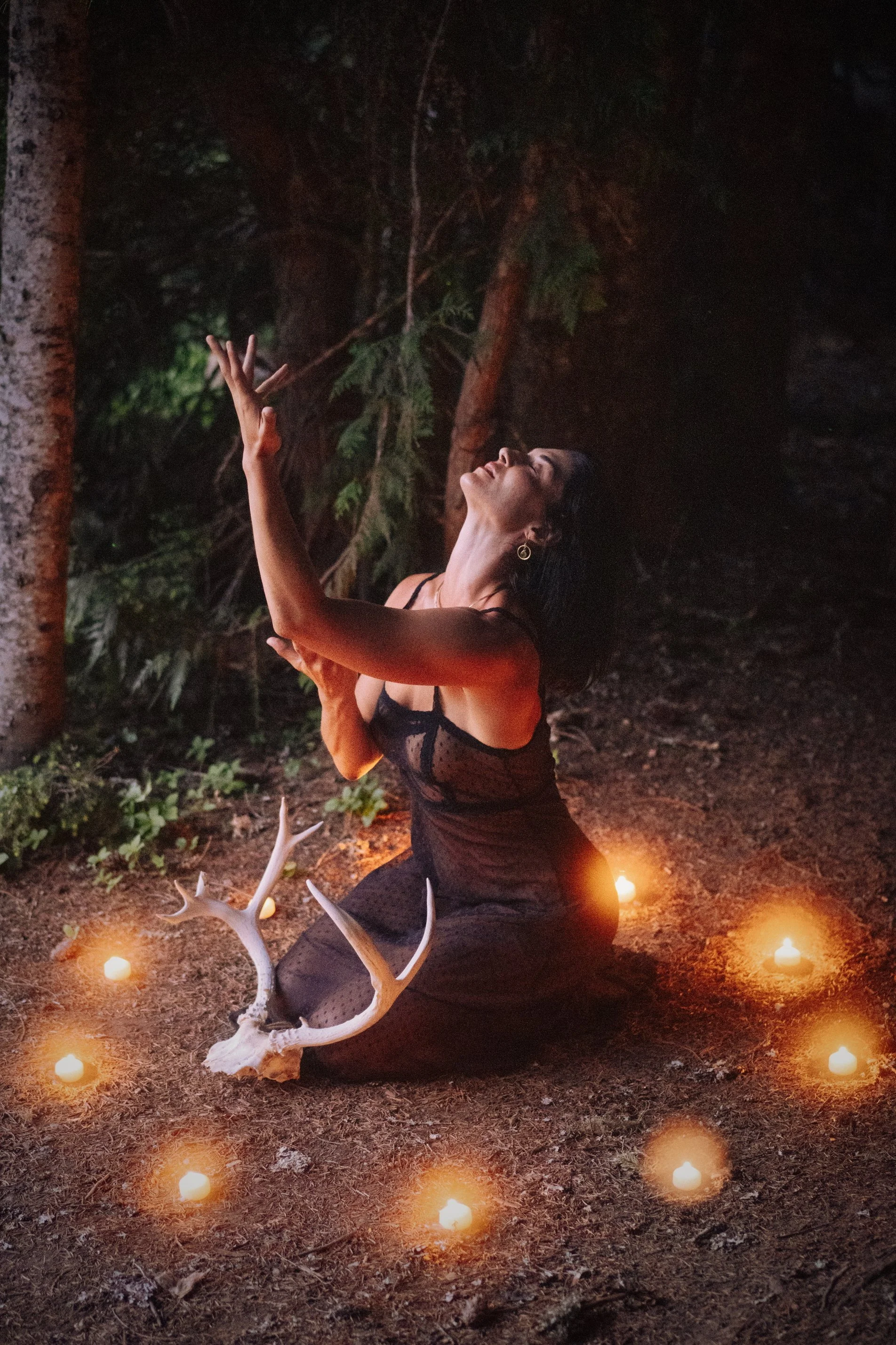 woman kneeling in a forest at night surrounded by candles and antlers with arms raised in a ritual pose