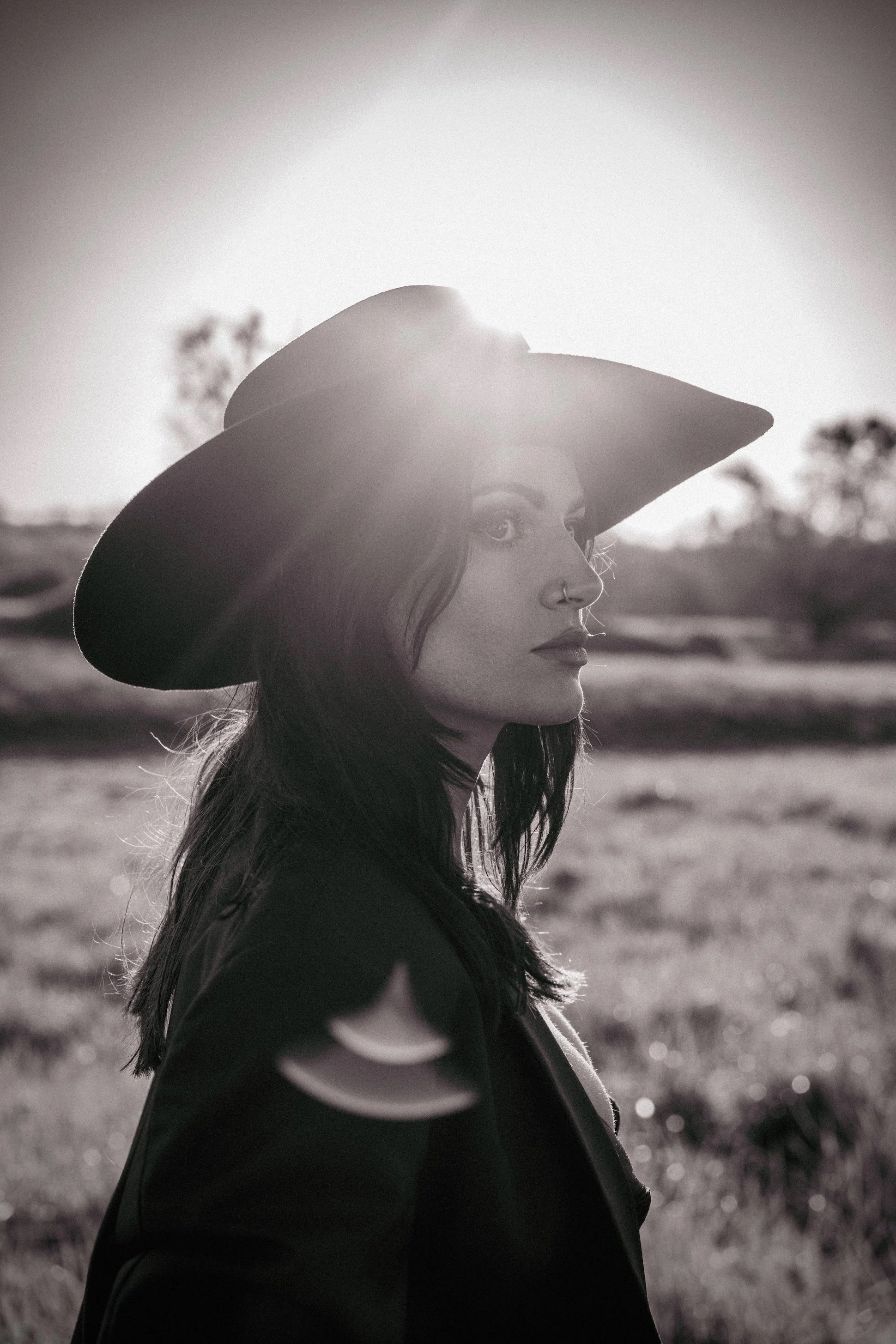 Black and white backlit portrait of woman in wide brim hat standing in a field with sun flare, fine art photography by Missoula Montana photographer