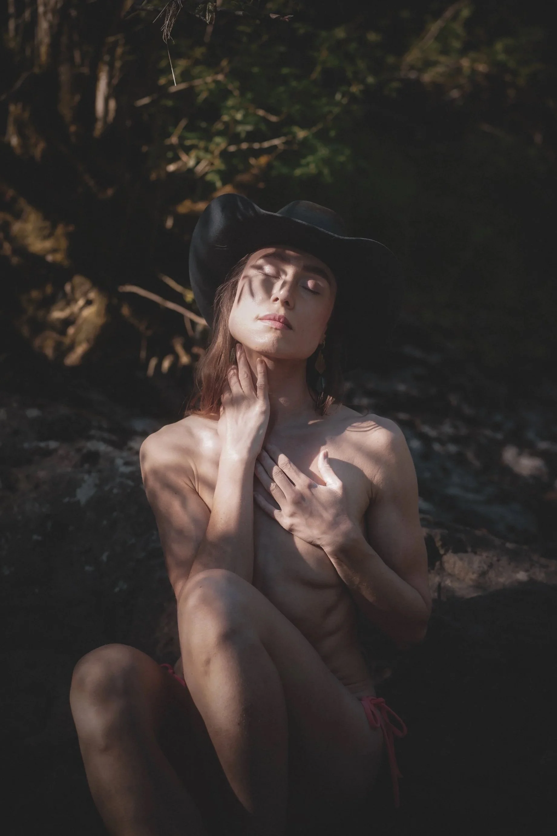 Woman with eyes closed sitting by a creek wearing a cowboy hat with dappled light across her face, intimate nature portrait photography by Montana photographer.