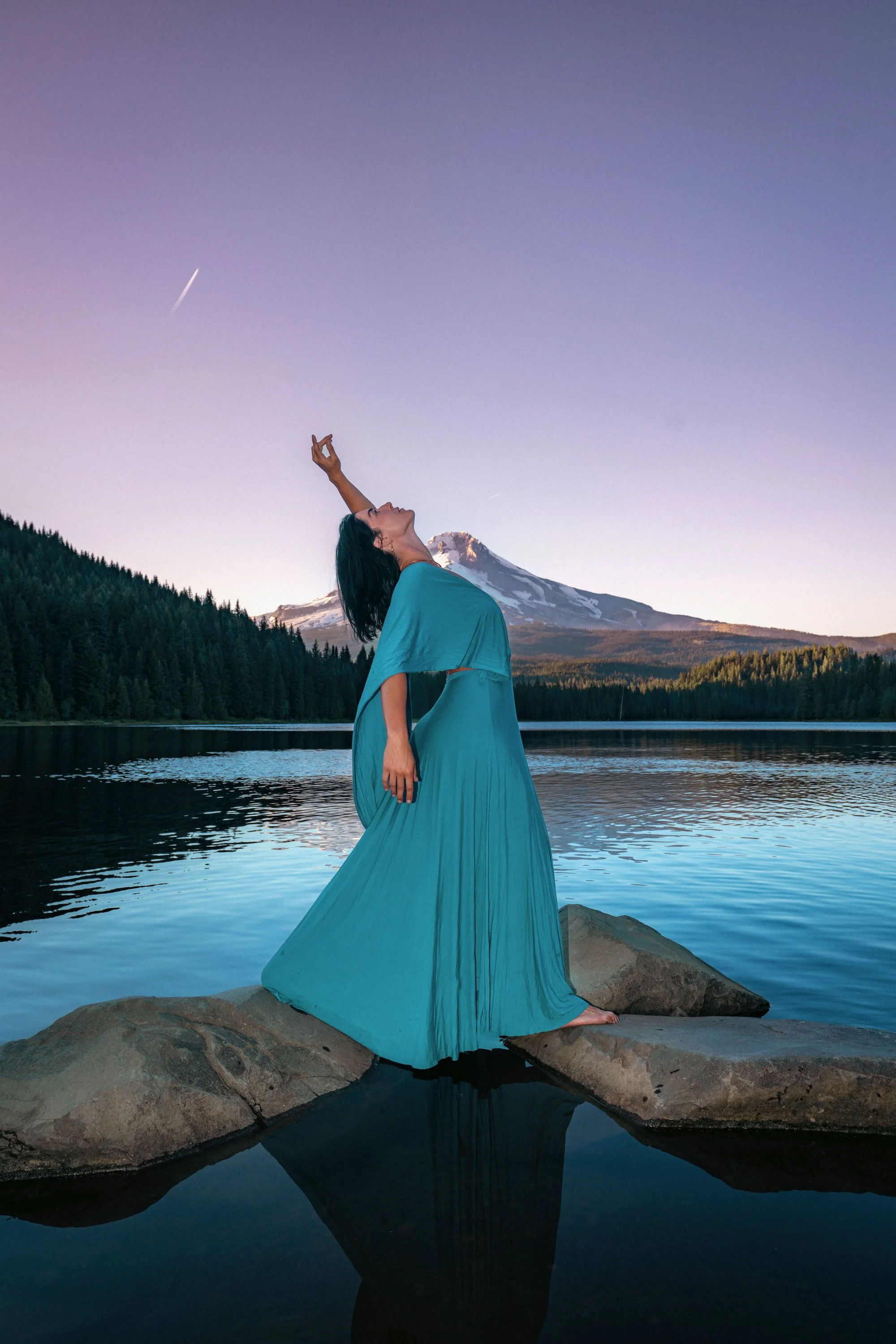 Woman in flowing teal dress standing on rocks at Trillium Lake reaching toward the sky at twilight with Mount Hood behind her, fine art nature portrait photography by PNW photographer