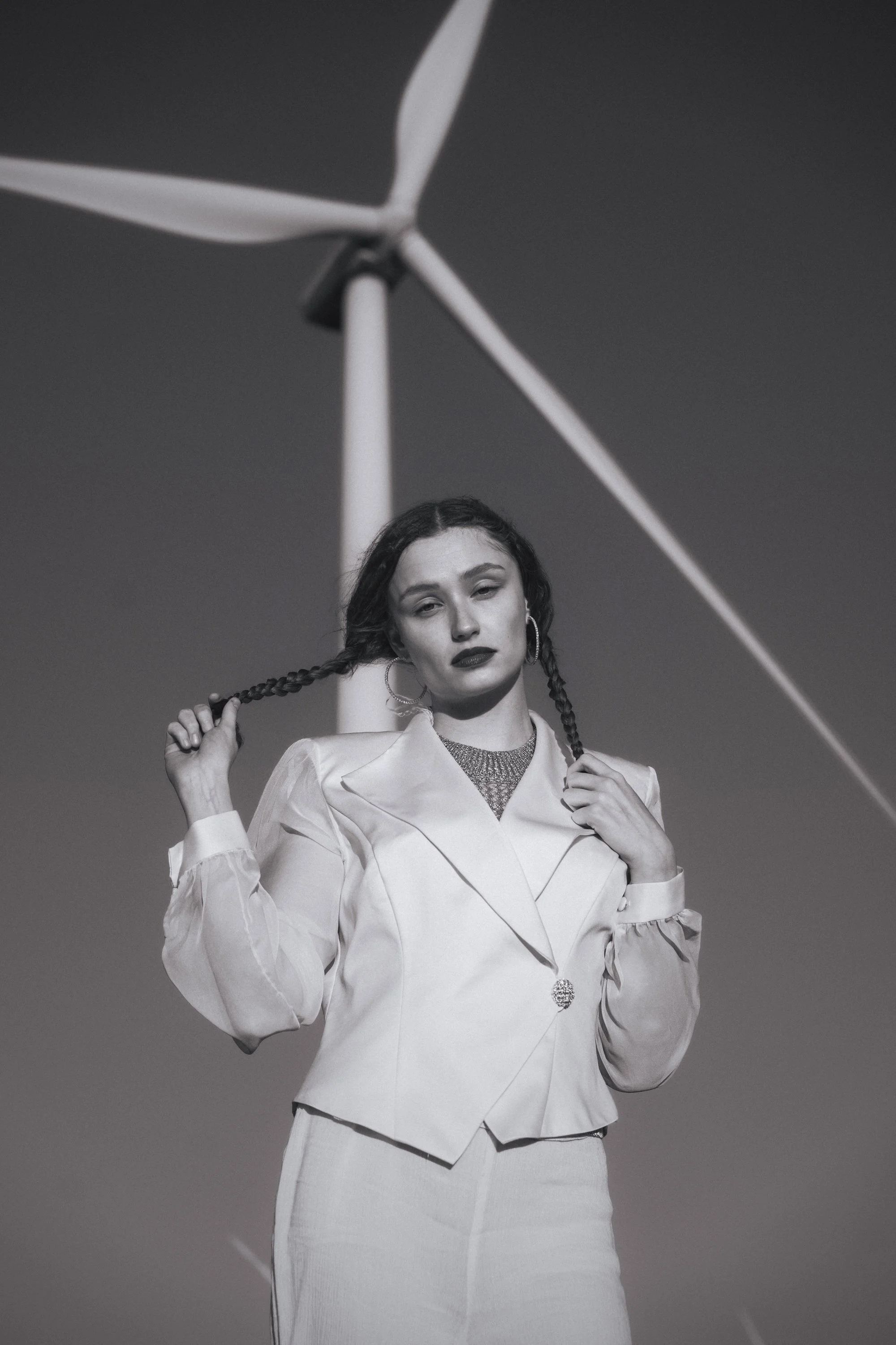 Black and white editorial portrait of woman in white suit with braids holding her hair beneath a wind turbine, bold conceptual photography by Montana photographer