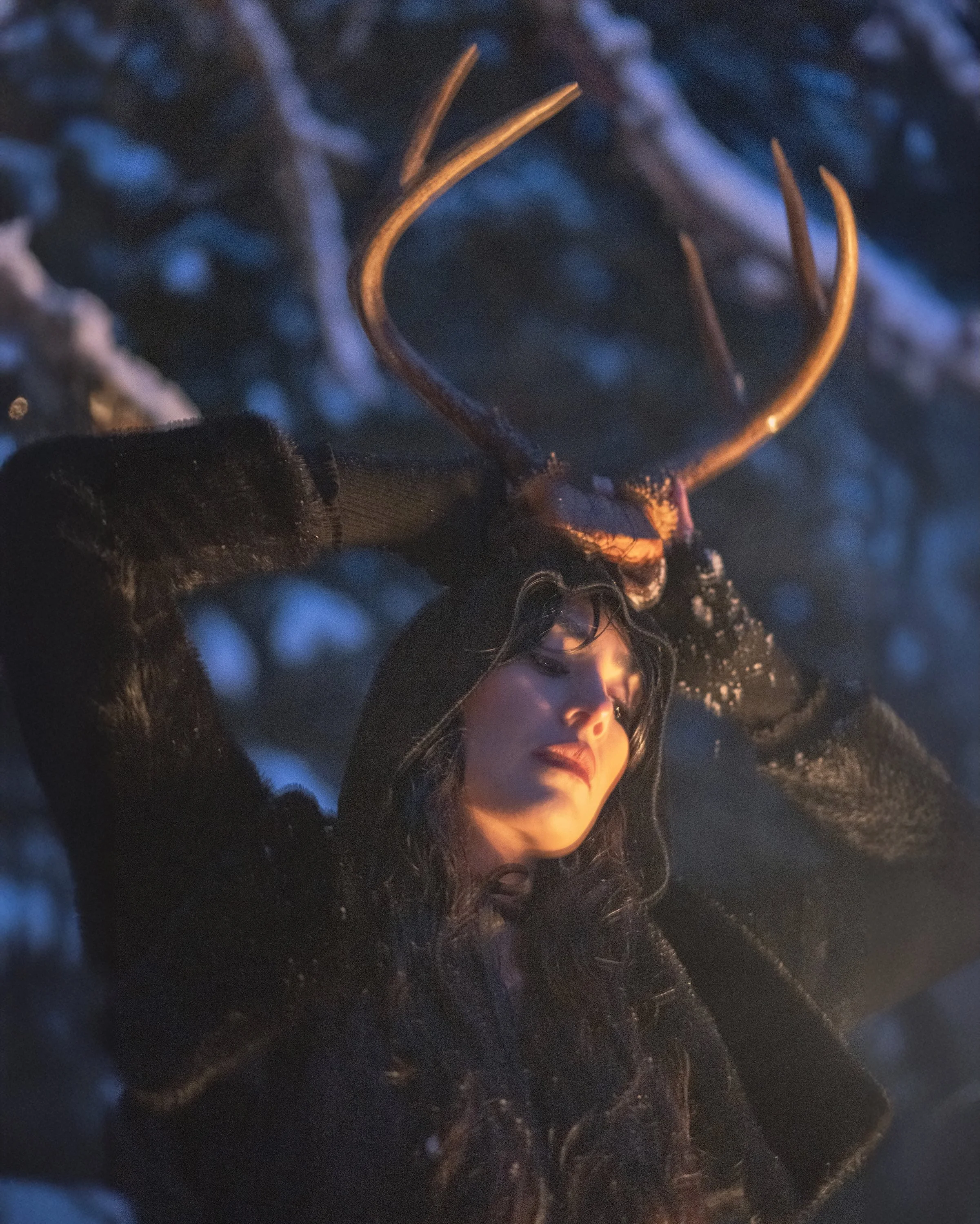 Woman holding antlers above her head in a snowy forest at night with warm golden light, ritual fine art portrait photography by Missoula Montana photographer