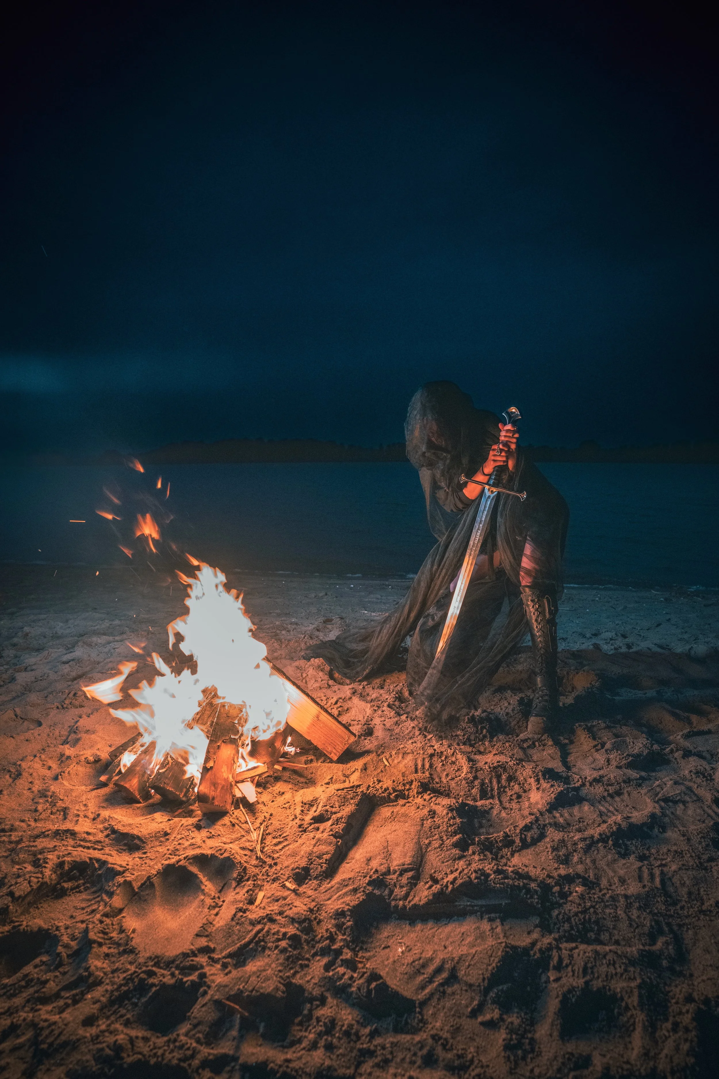 Hooded woman holding a sword beside a bonfire on a dark beach at night, conceptual fine art portrait photography by Montana photographer
