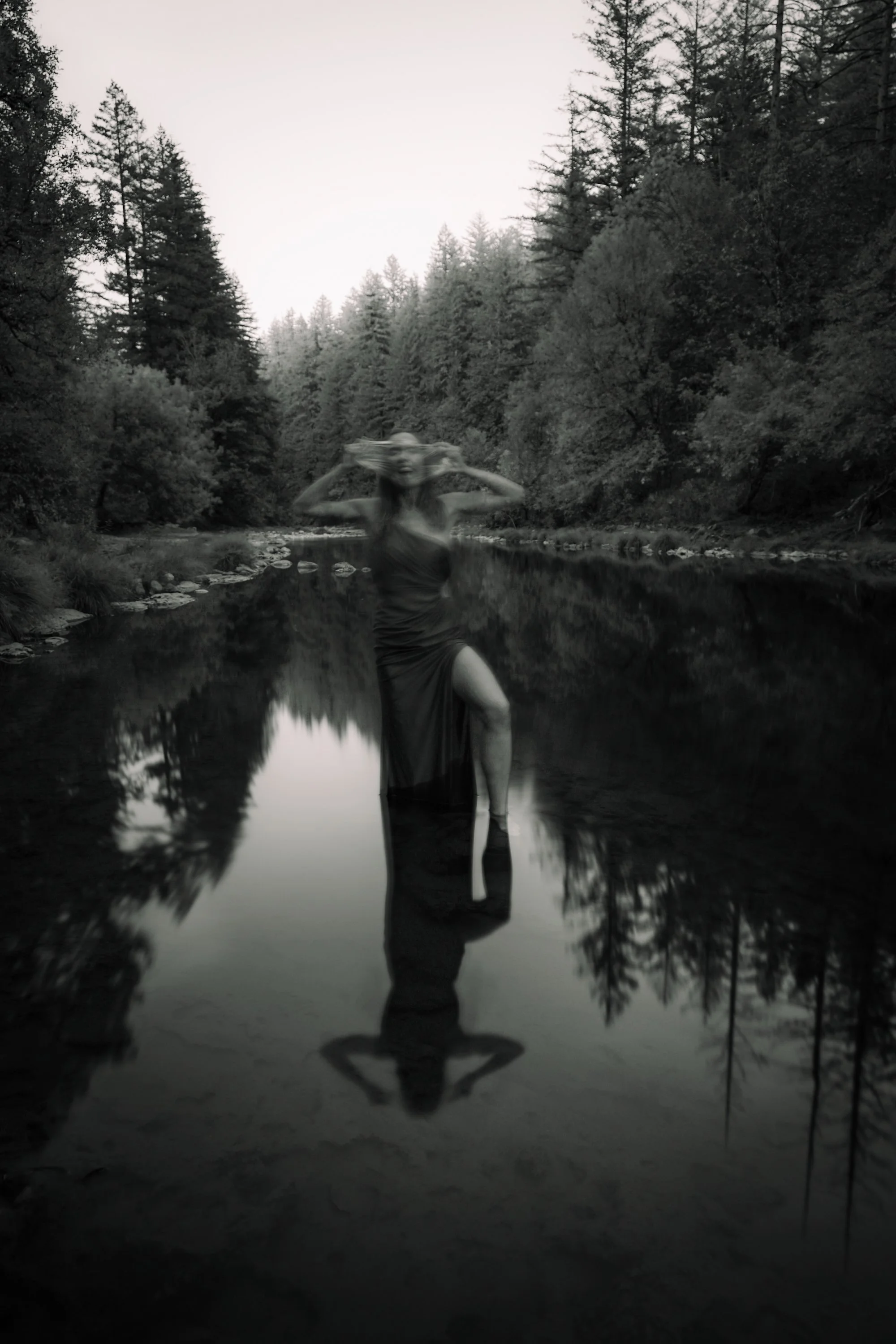 Black and white self portrait of woman standing in a river with full reflection in still water surrounded by forest trees, nature portrait photography by Montana photographer