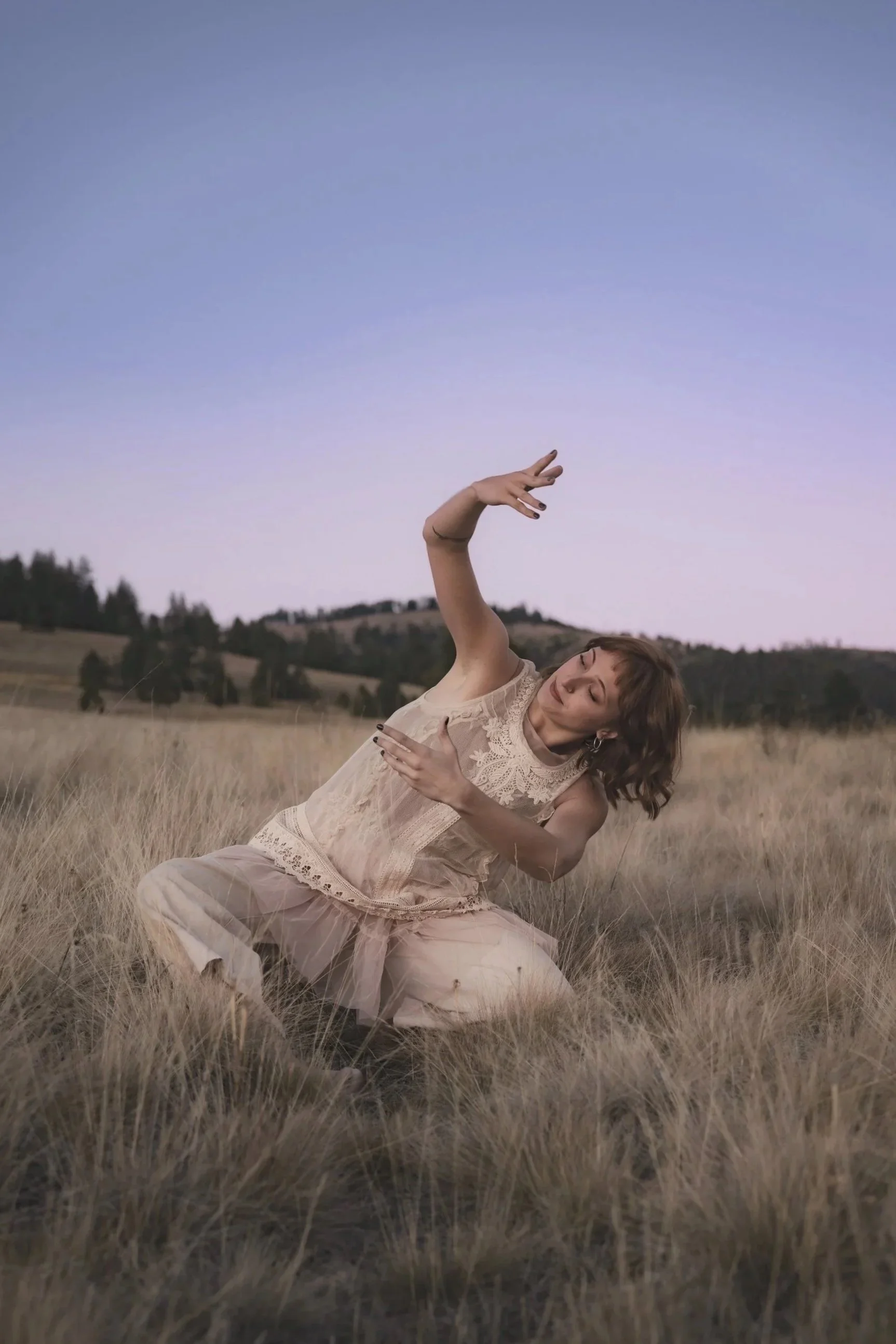 Woman in vintage lace dress dancing in dry grass field at twilight with Mt Sentinel and purple sky behind her, wild nature portrait photography by Missoula Montana photographer.