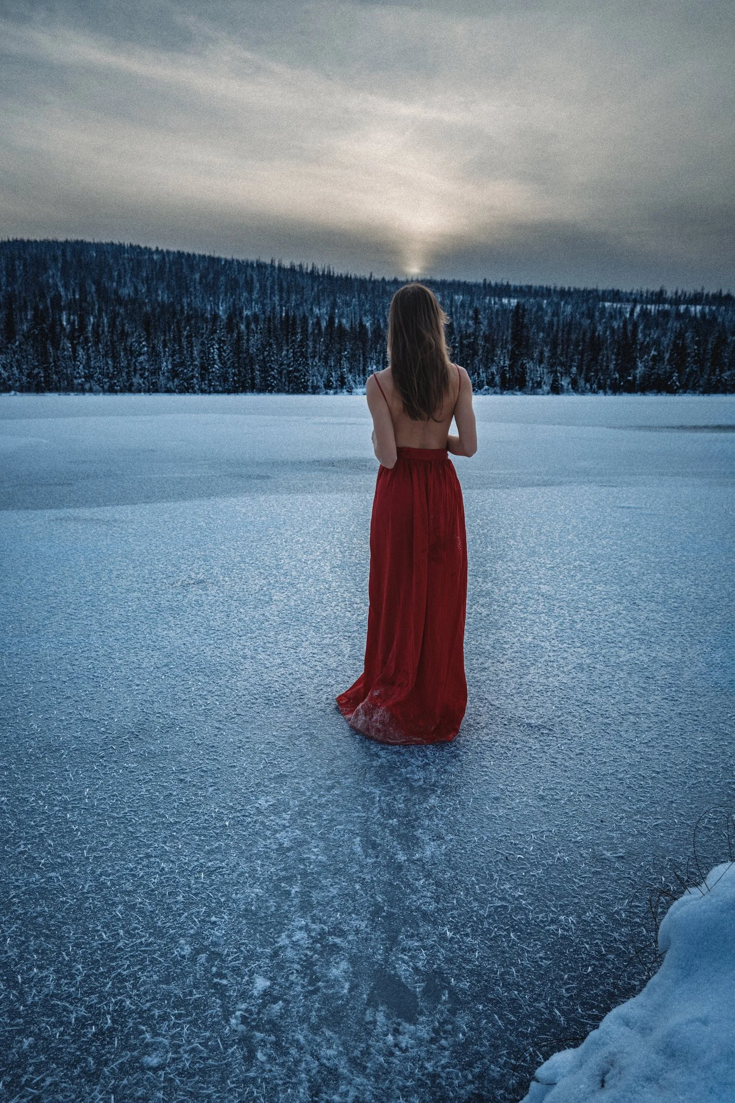 Self portrait of woman in red dress standing alone on a frozen lake at winter sunset facing the horizon with snow covered forest behind her, fine art empowerment photography by Missoula Montana photographer.