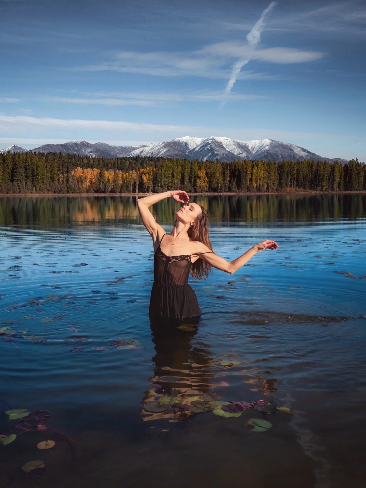 Self portrait of woman in black dress standing in Seeley Lake Montana with snow capped mountains and autumn forest reflected behind her, wild nature photography by Missoula Montana photographer
