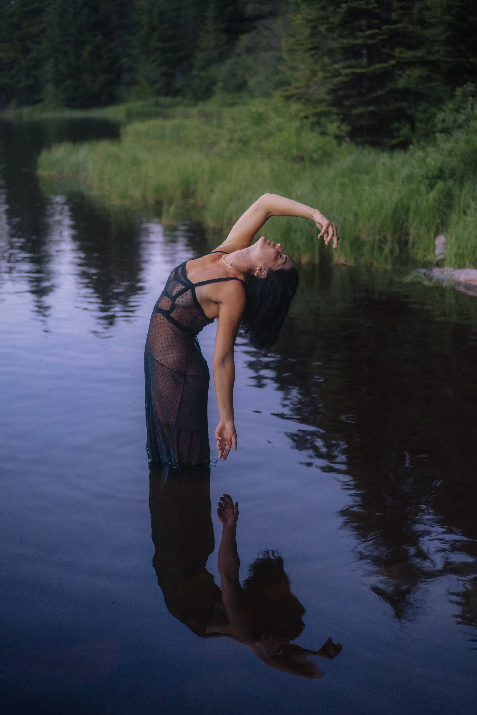 Woman in sheer black dress arching freely in Seeley Lake Montana with full reflection in still water, female empowerment fine art nature photography by Missoula Montana photographer