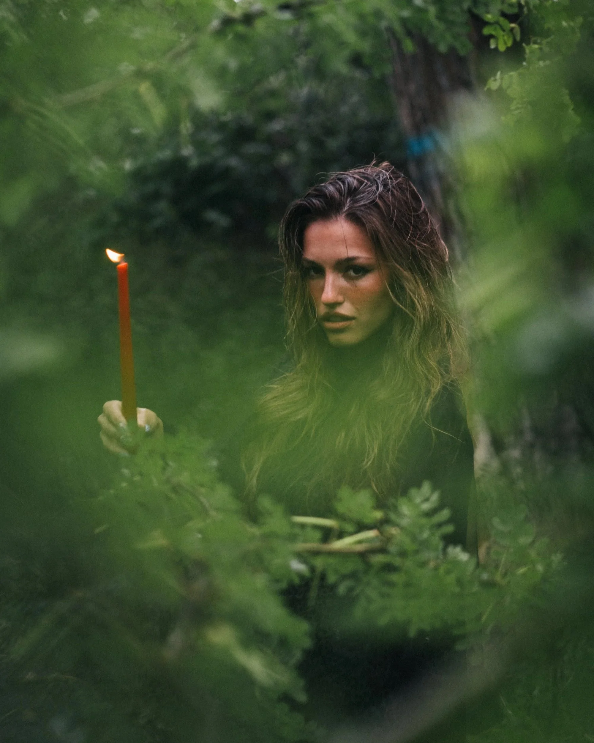Woman holding a lit candle framed through lush green foliage in a forest, wild nature portrait photography by Missoula Montana photographer