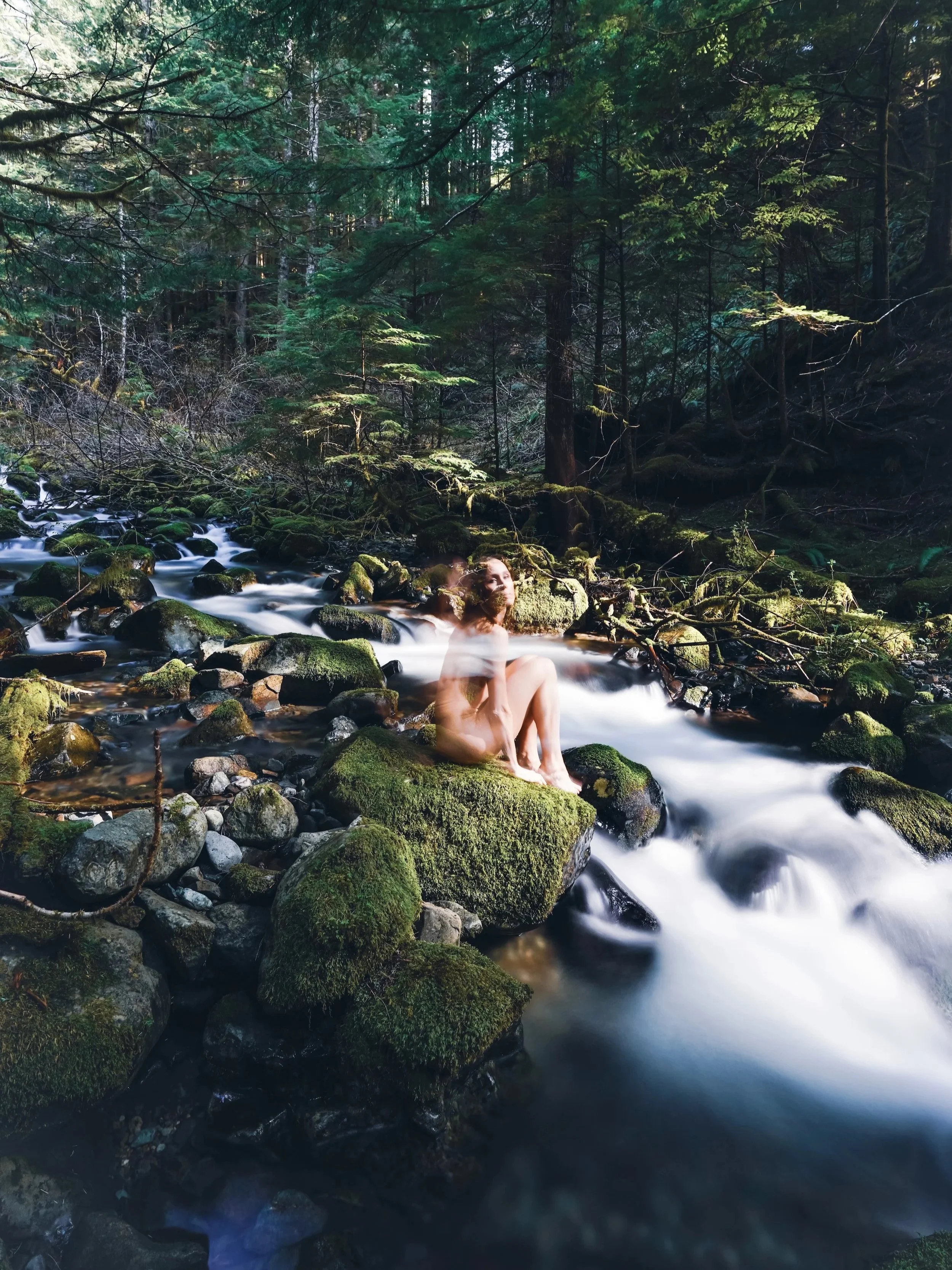 Long exposure self portrait of woman sitting on mossy rocks in PNW creek surrounded by flowing water and pine forest, fine art nature photography by Missoula Montana photographer