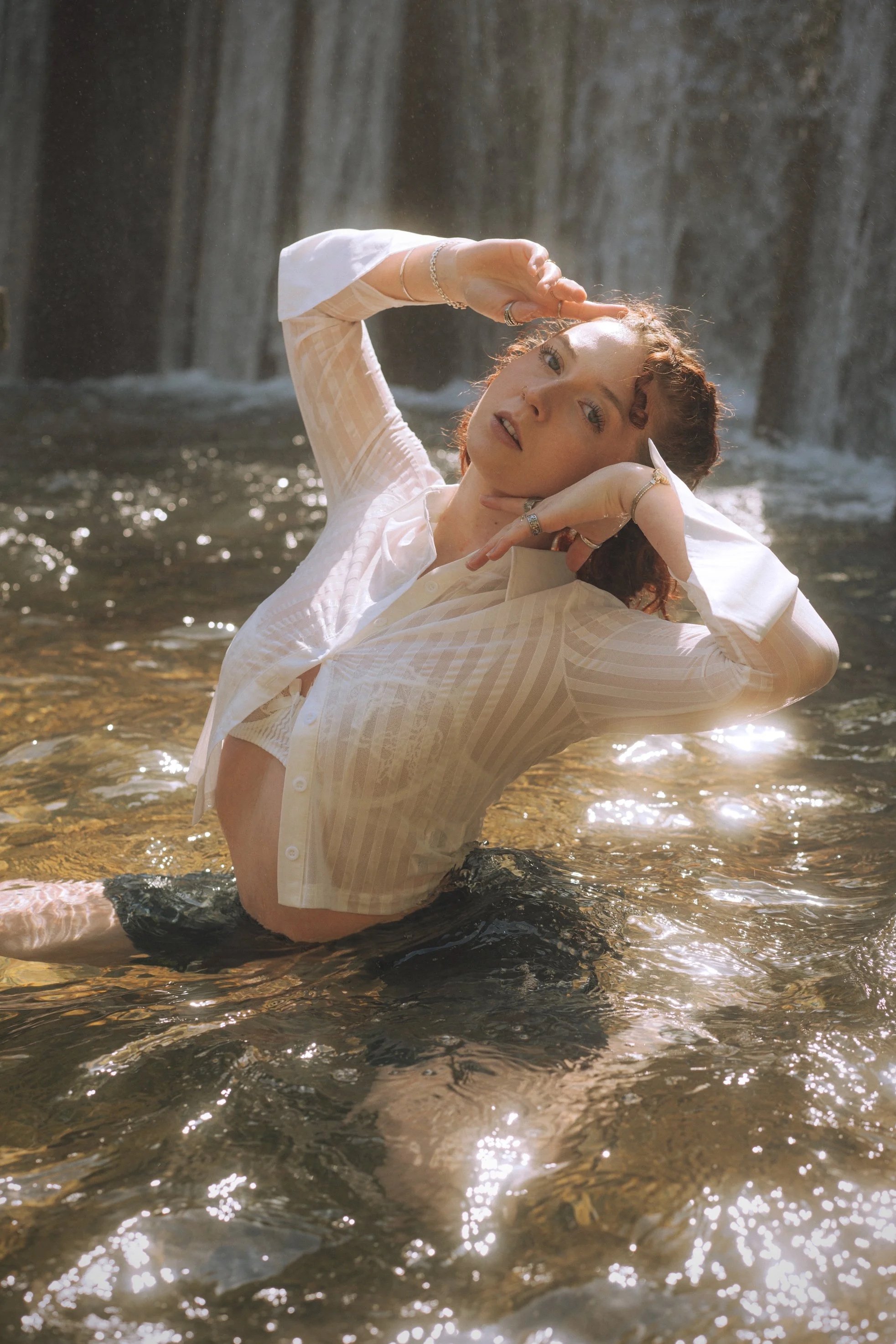 A woman with curly red hair in a white semi-transparent shirt, partially submerged in a shallow creek or river, with a waterfall in the background. She is posing with one hand on her head and the other near her face, surrounded by sunlight reflecting off the water.