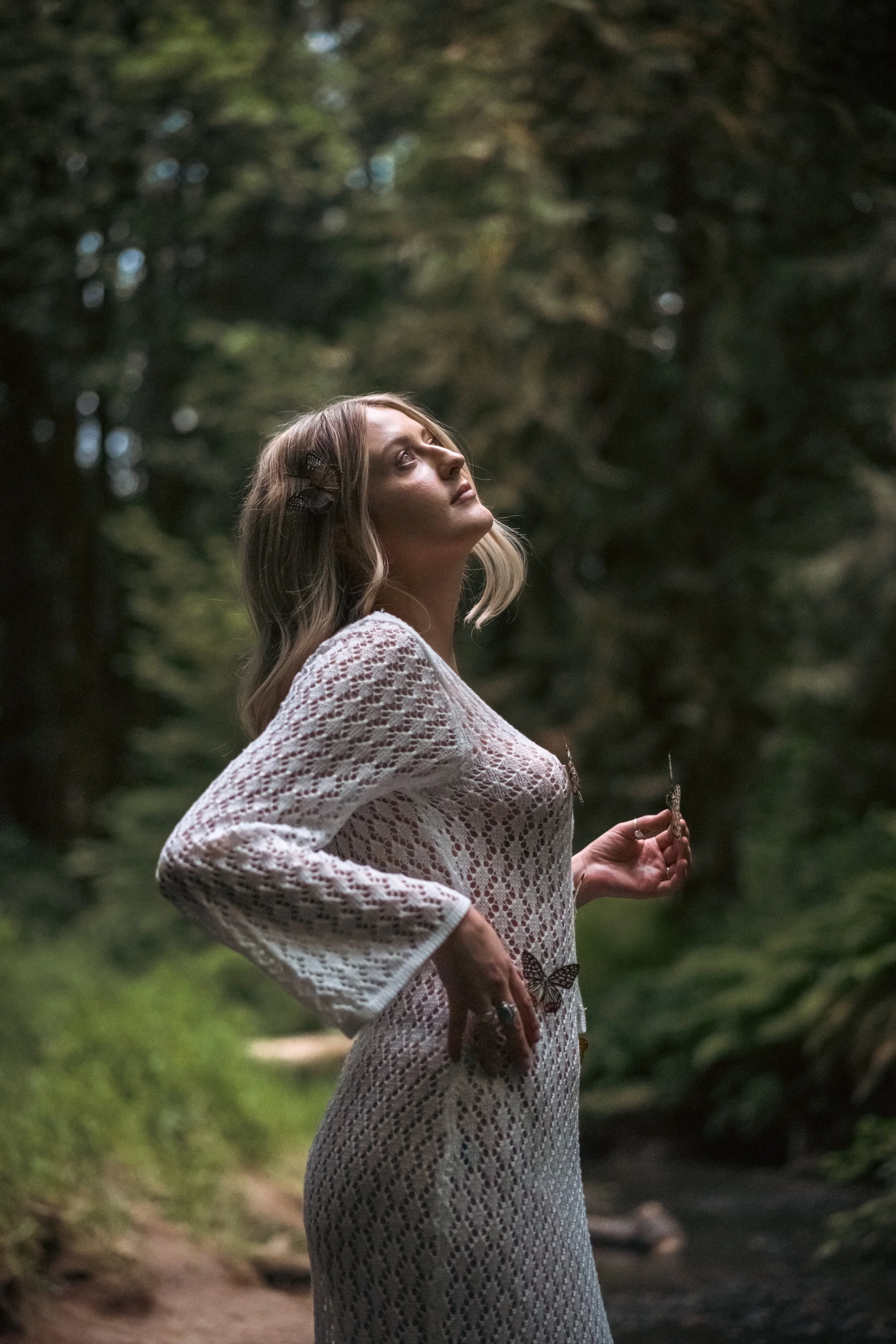 Woman in white lace dress with butterflies on her body looking up toward the sky in a pine forest, ethereal fine art portrait photography by Missoula Montana photographer.