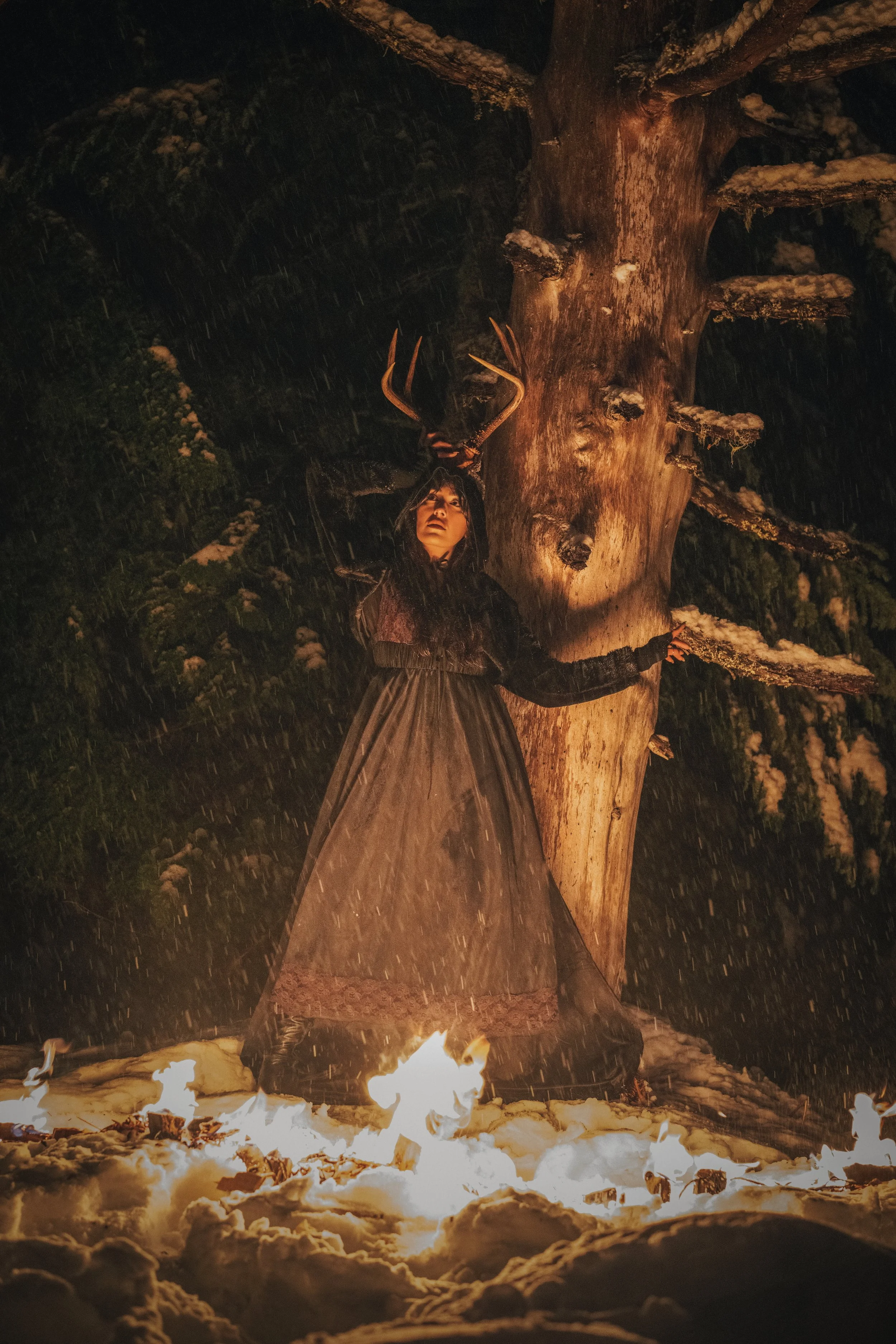 Woman wearing antlers in a dark snowy forest standing beside a tree with fire burning at her feet in the rain, bold ritual fine art portrait photography by Missoula Montana photographer.