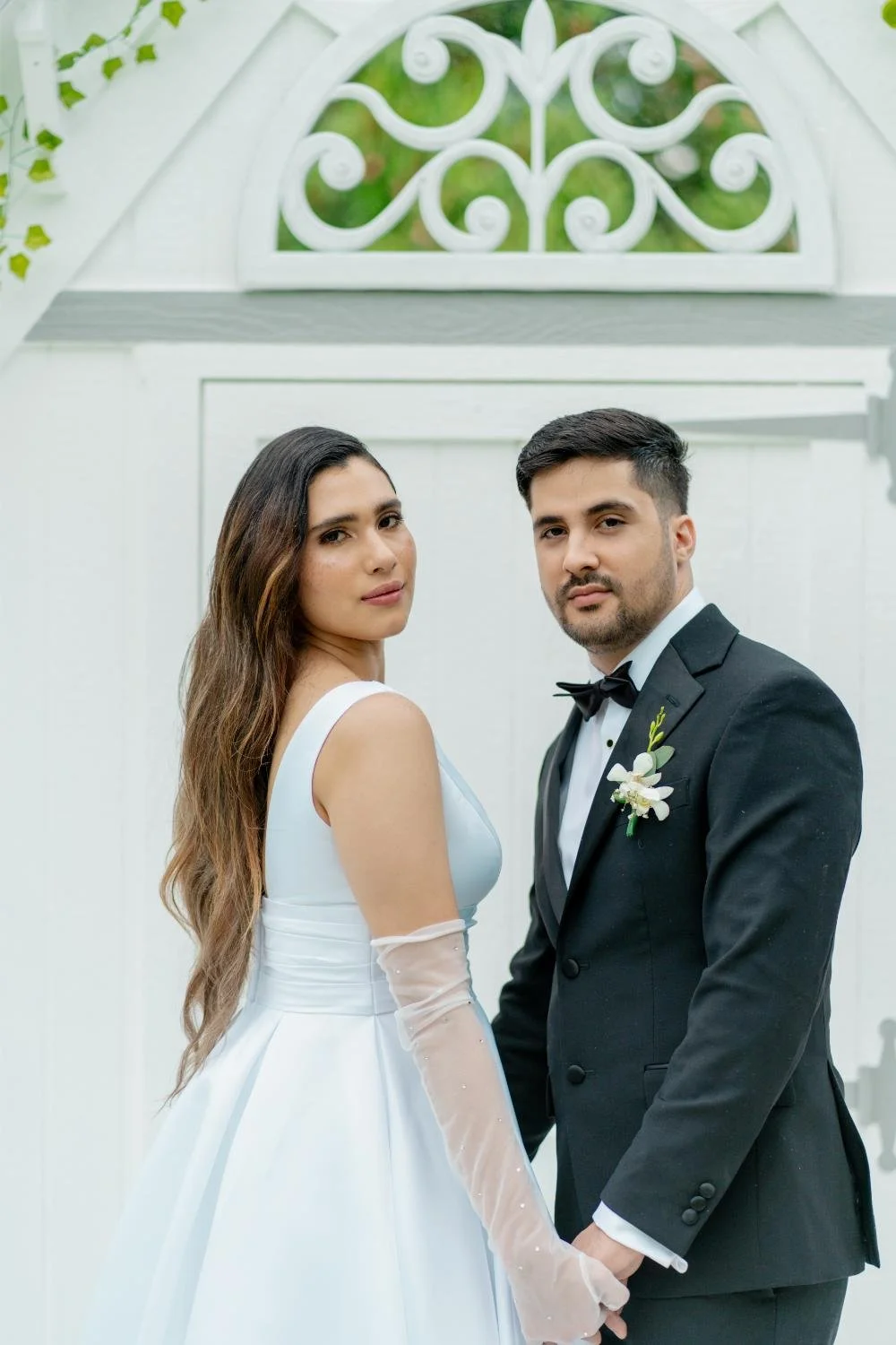 A bride and groom holding hands and standing in front of a white wall with a decorative window, dressed in wedding attire with the bride in a white gown and the groom in a black tuxedo, outdoors during daytime.