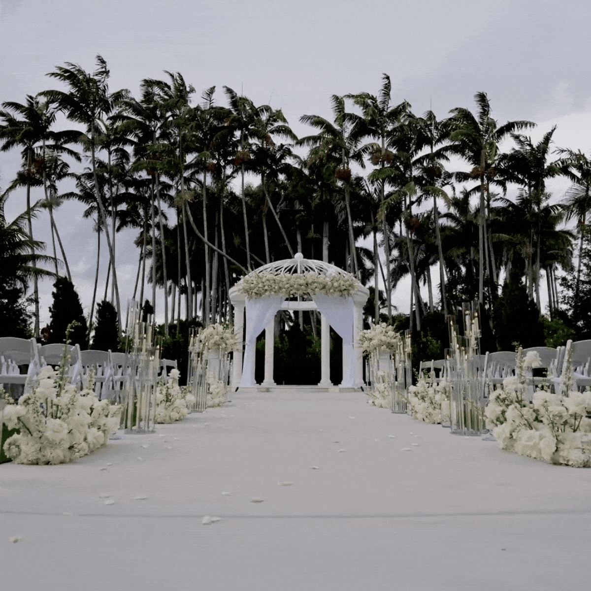 Outdoor wedding altar decorated with white flowers and drapes, set in front of tall palm trees on an overcast day.