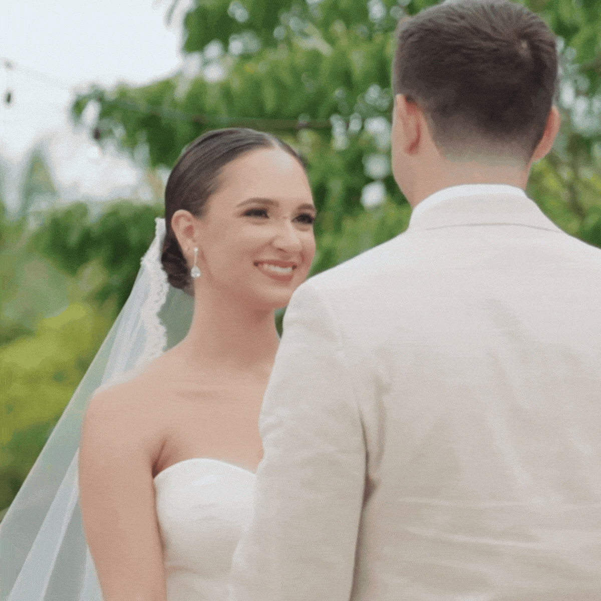 A bride and groom exchanging vows outdoors, with the bride smiling and looking at the groom, surrounded by greenery.