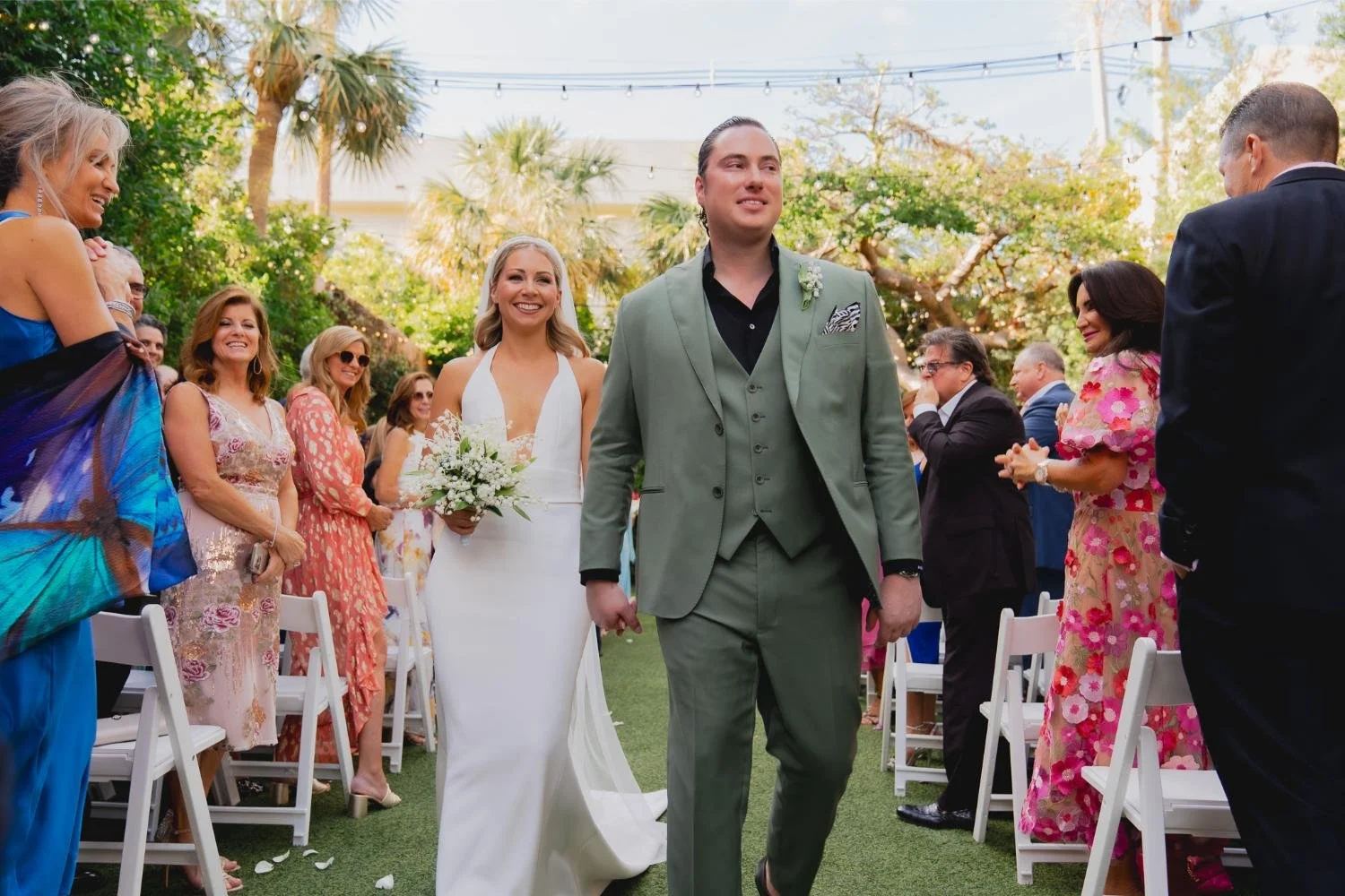 A wedding ceremony outdoors with a bride in a white dress holding a bouquet, walking down the aisle with a groom in a green suit, surrounded by guests clapping and smiling in a garden setting with trees and string lights.