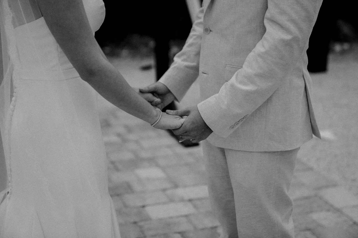 A couple holding hands during a wedding ceremony, with the bride in a wedding dress and the groom in a light suit, standing on cobblestone pavement.