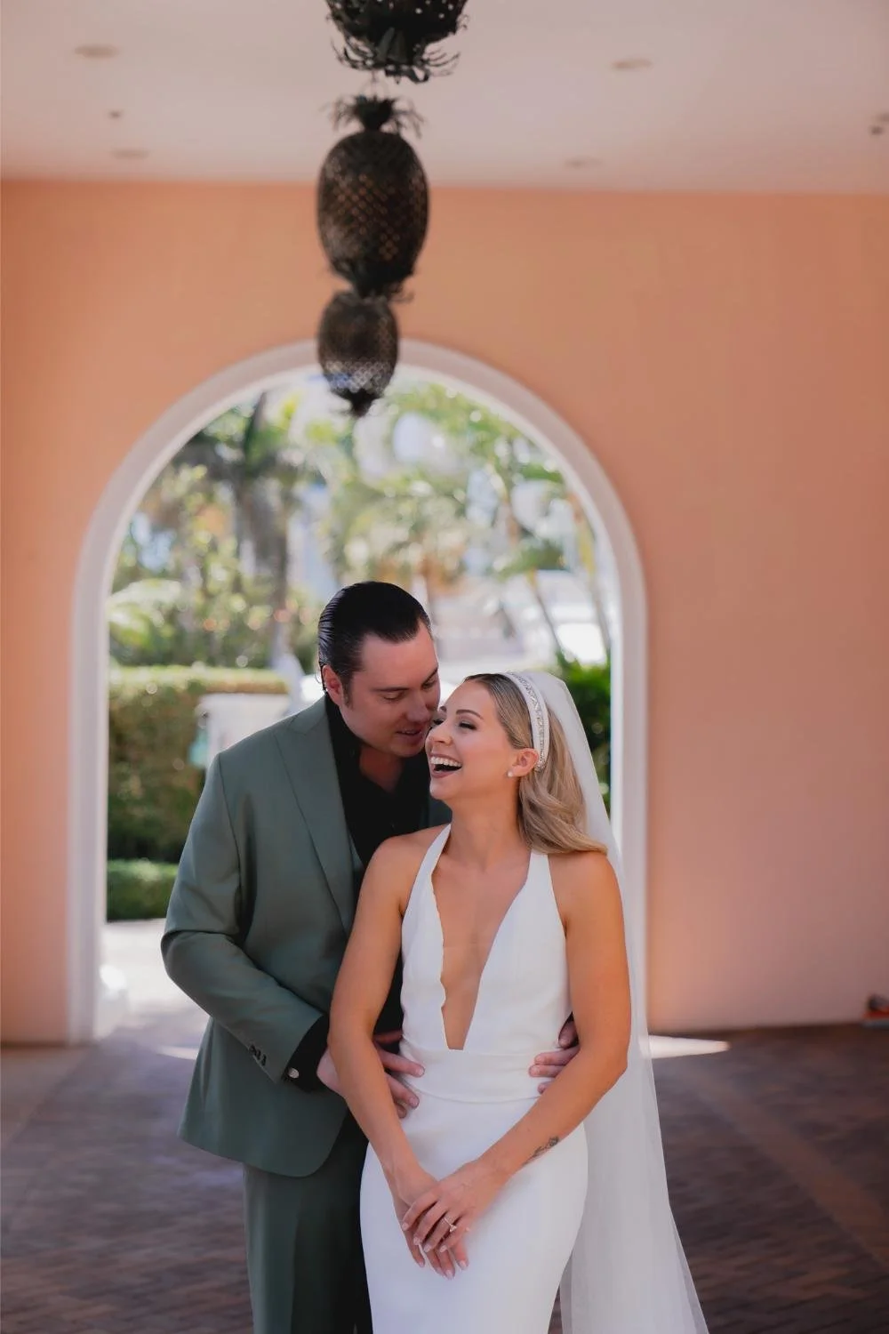 A happy couple dressed in wedding attire, standing close together inside a room with a large arched window and outdoor greenery in the background.