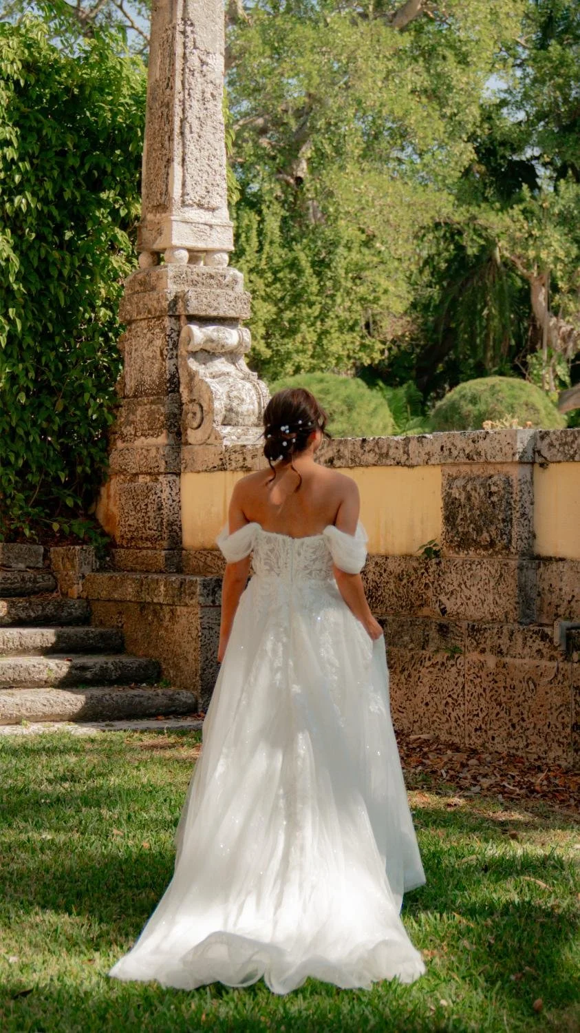A woman in a white wedding dress with off-the-shoulder sleeves stands outdoors in front of old stone architecture, surrounded by green trees and grass.