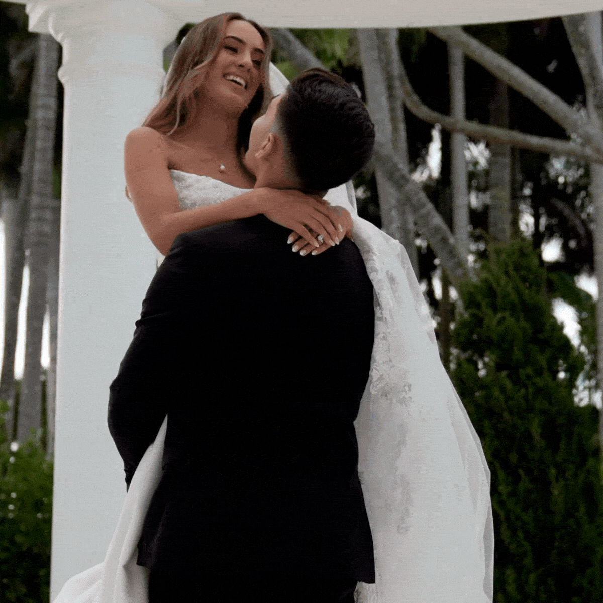A bride and groom embrace during their wedding ceremony outdoors, with trees and a white arch in the background.