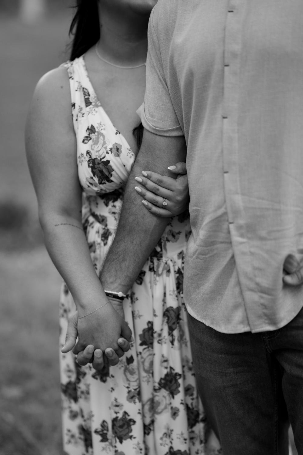 A close-up of a couple holding hands, with the woman wearing a floral dress and an engagement ring, and the man dressed in a casual, short-sleeved shirt.