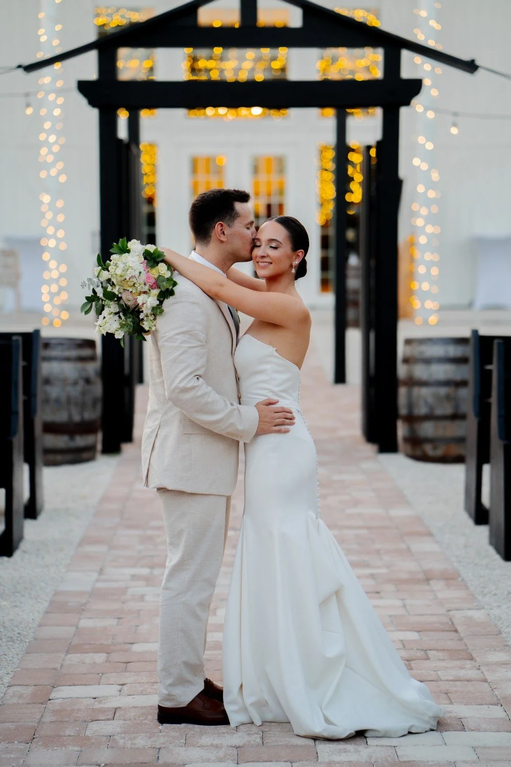 A bride and groom sharing a kiss outdoors during their wedding, with the bride holding a bouquet of flowers. The background features string lights and a decorative arch.