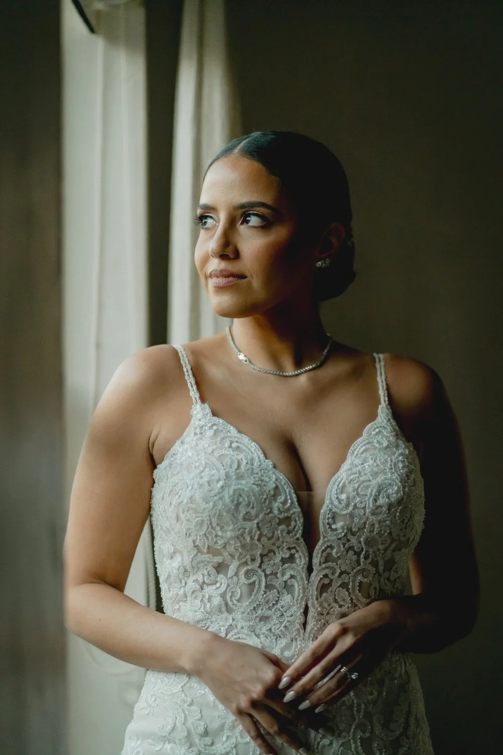 A woman in a white lace dress standing by a window, looking outside with a thoughtful expression.