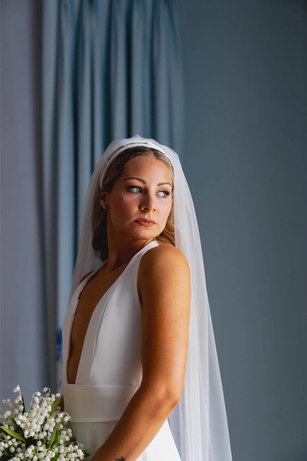 Bride in a white dress and veil holding a bouquet of white flowers stands near a blue wall and curtain, looking to the side with a contemplative expression.