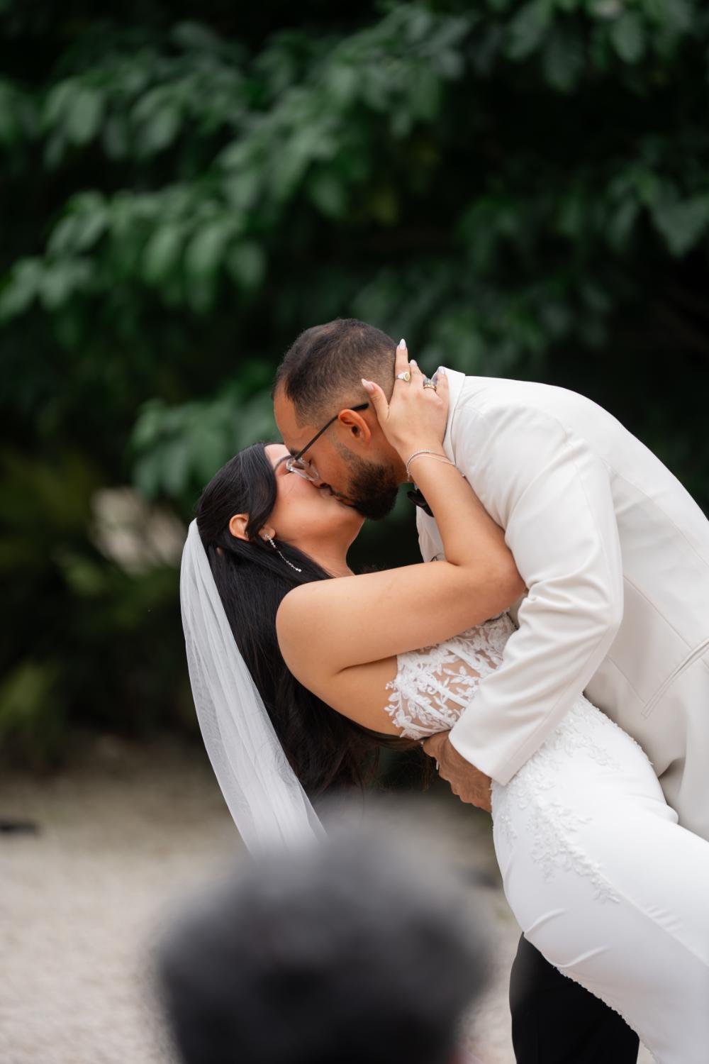 A bride and groom share a kiss outside, with the groom dipping the bride. The bride is wearing a white wedding dress with lace details and a veil, while the groom is wearing a white suit. Green foliage is visible in the background.
