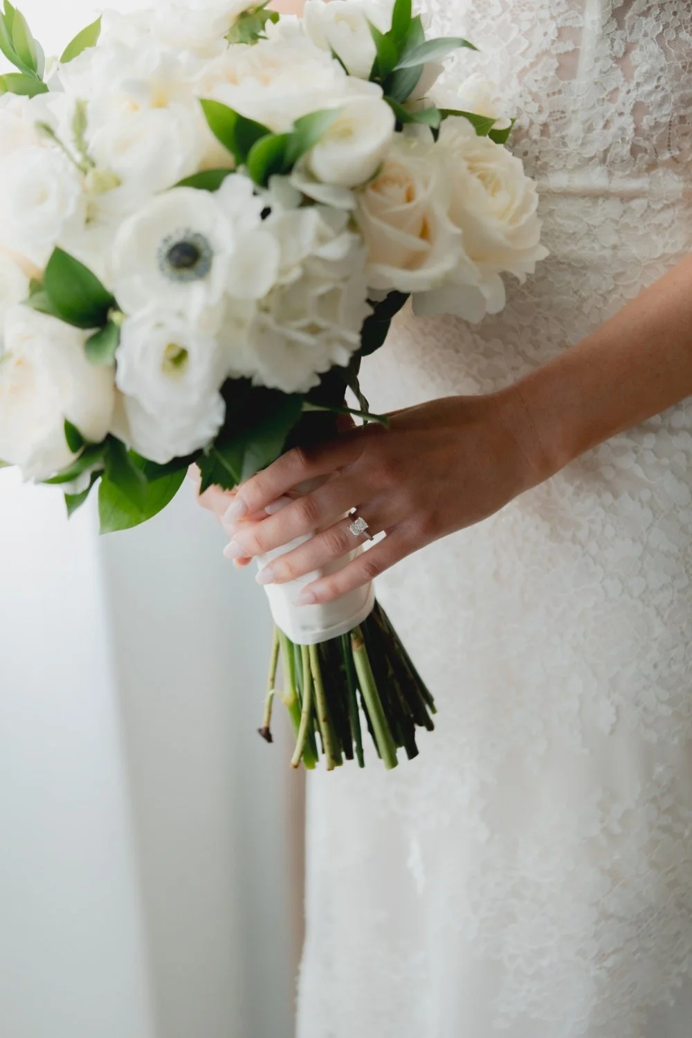 A bride holding a bouquet of white roses, anemones, and greenery, with her left hand showing a diamond engagement ring.
