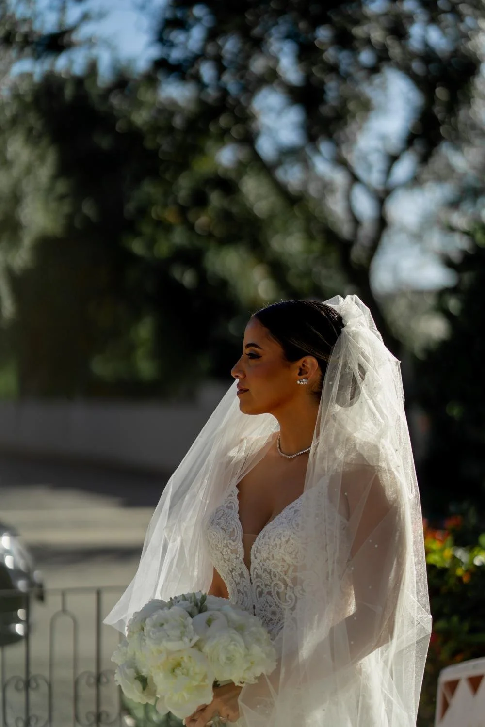 Bride with dark hair in an elegant wedding dress, holding a bouquet of white flowers, standing outdoors with a blurred background of trees and sky.